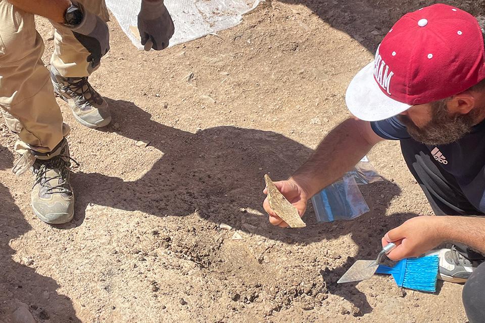 A team member holds a piece of brick or stone in one hand and an archeological implement in the other. The dusty, booted feet of a colleague are seen on the side of the image.