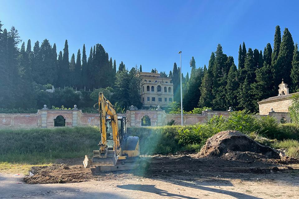 A piece of earth-moving equipment sits on a pile of dirt, with an ornate wall and home on a hillside in the background.