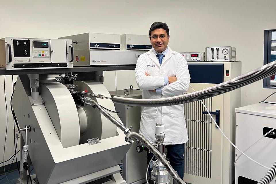 Reza Dastvan, Ph.D., in his lab, wearing a lab coat and standing next to some large pieces of equipment.