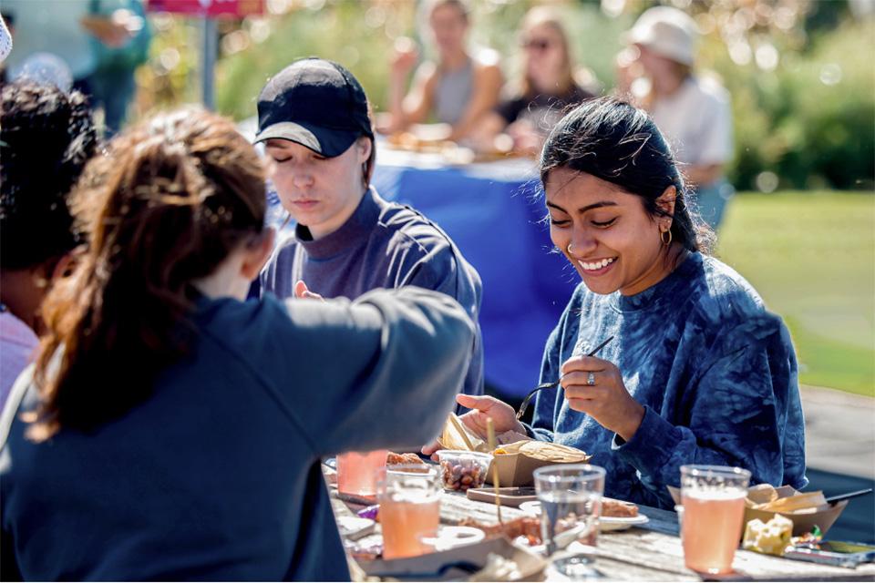 博彩网址大全 students sit at a picnic table filled with cups and cardboard trays filled with food on a sunny day.