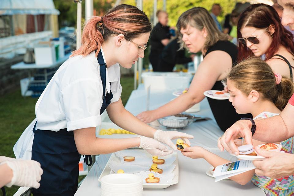 A student wearing an apron reaches over a table to hand a piece of food to a child.