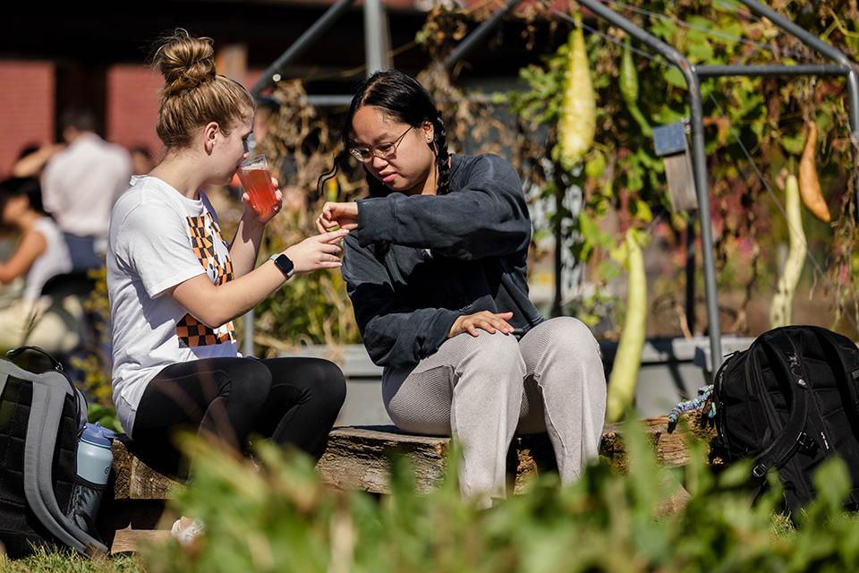 Two students sit on a bench outdoors. One student sips on a beverage while holding up a piece of food to share with her neighbor.