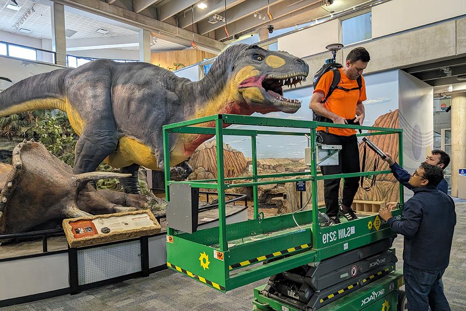 Cagri Gul, master's student in geographic information science, gets situated with the LiDAR backpack scanner before beginning to scan the repaired T. rex. Also pictured: Alifu Haireti, geospatial computing engineer with TGI, and Mustafizur Rahaman, master's student in geographic information science.