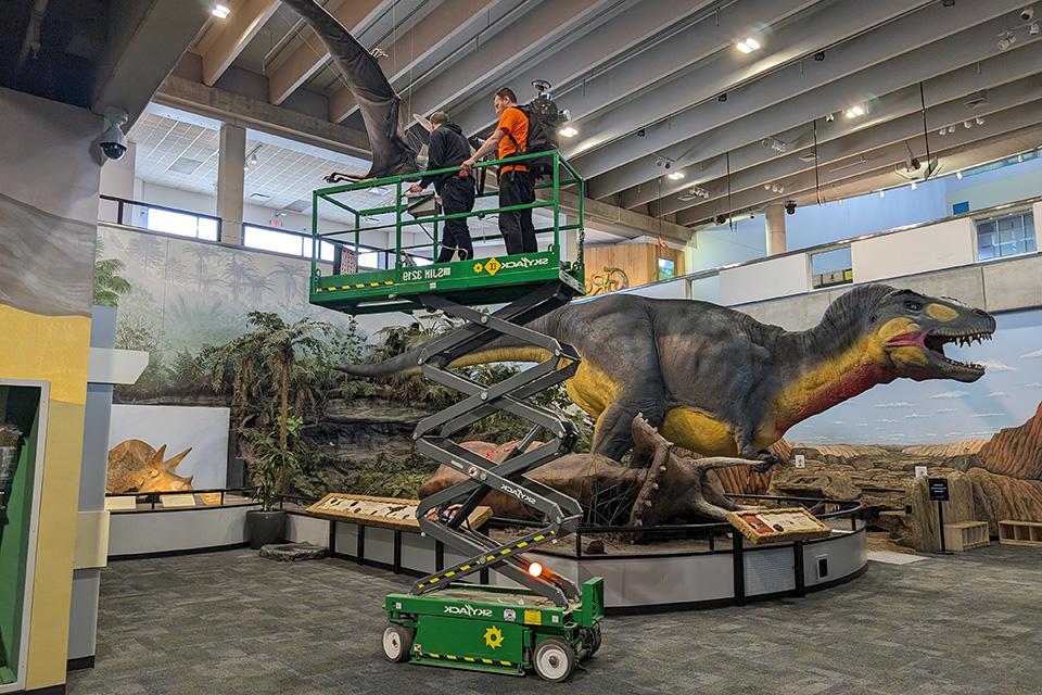 Cagri Gul, master’s student in geographic information science, stands atop the SkyJack as Chris Lucas, exhibit designer with the Saint Louis Science Center, drives. The scanning team needs to capture angles of the diorama from above in order to create a more complete scan; simply walking with the backpack on the ground would not collect data from the top of the dinosaurs.