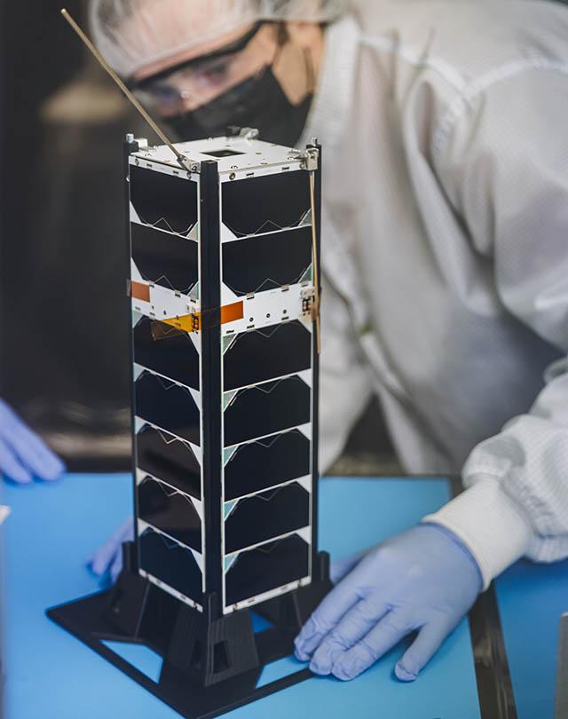 A small satellite with a height of about about 30 centimeters long and 10 centimeters on its side shaped like a metal loaf of bread sits on a table. A student wearing protective gear is seen in the background.