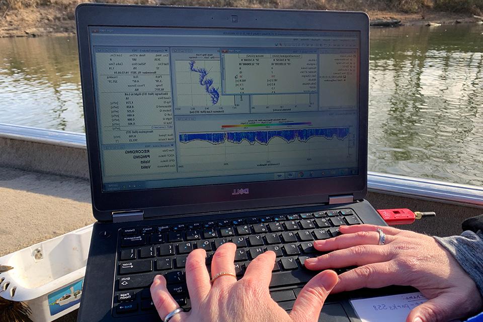 A pair of hands type on a laptop keyboard, while the screen shows charts, graphs and data sets, situated with a river in the background.