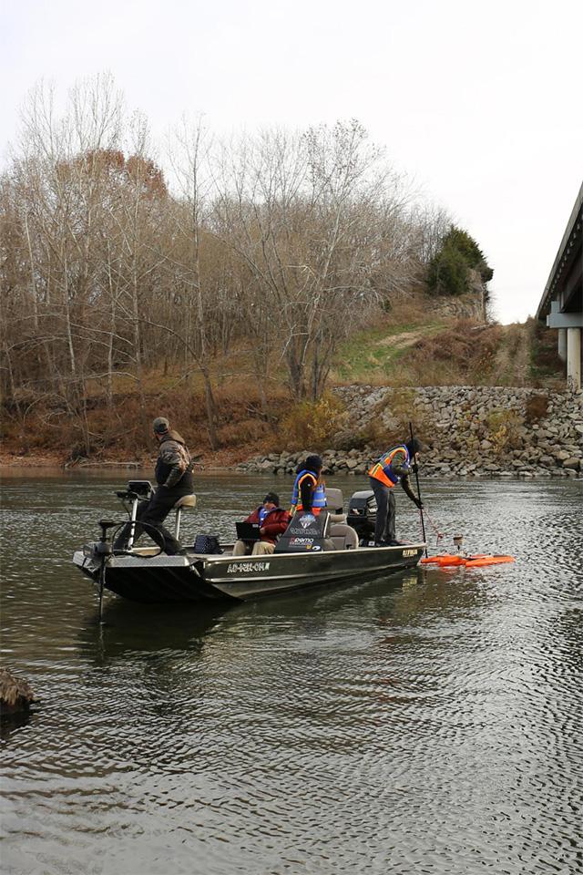 Four people are on a boat in a river, working with various pieces of equipment. Behind them, a shoreline with rocks and trees without leaves are visible.