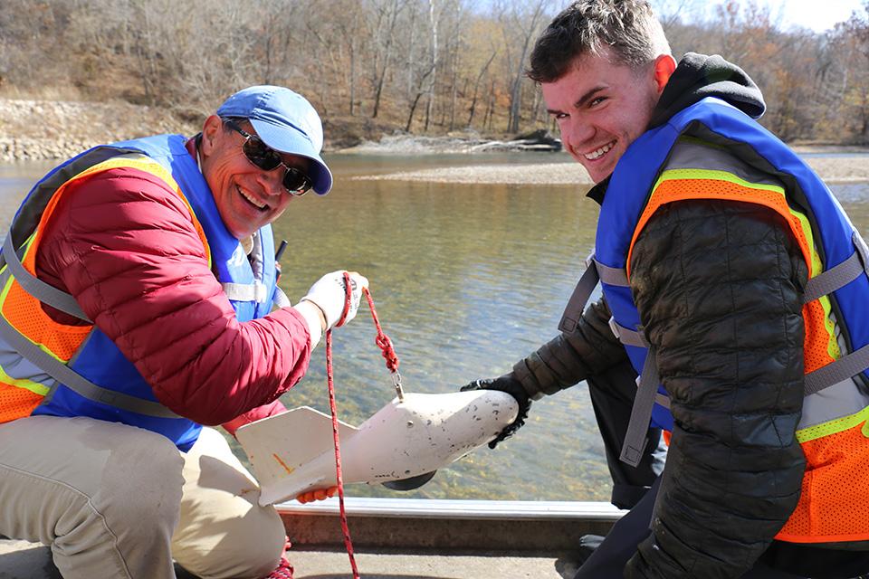 Two men wearing life jackets and reflective gear sit in a small boat on a river. They are holding a small piece of equipment on a rope. Rocky shore line with bare trees is visible in the background.